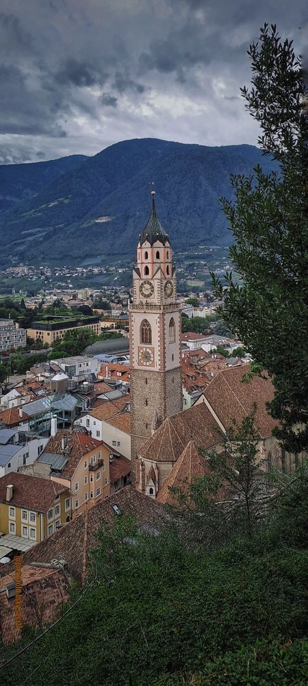 Panorama sul centro storico di Merano (Bolzano) dalla Tappeinerweg. Al centro della foto spicca il caratteristico campanile del Duomo di San Nicolò, di cui si vede anche la parte absidale. Sullo sfondo il Vigiljoch (Monte San Vigilio) sovrastato da nuvole cariche di pioggia 