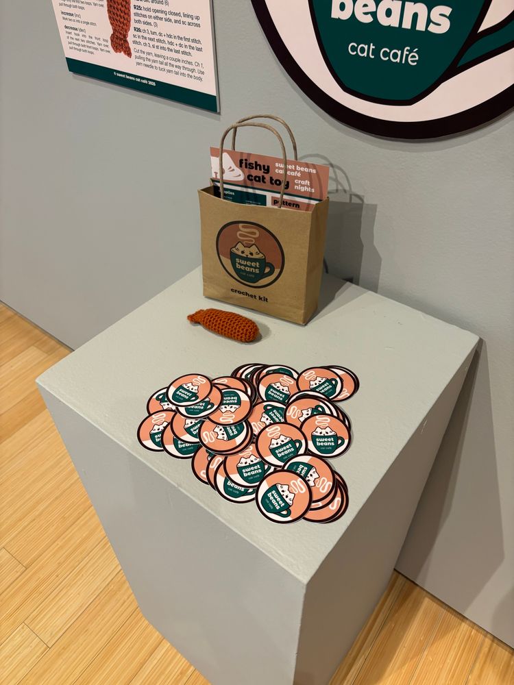 a photograph of just the podium, with the branded brown paper bag, a crochet fish, and a pile of stickers of the cafe’s logo