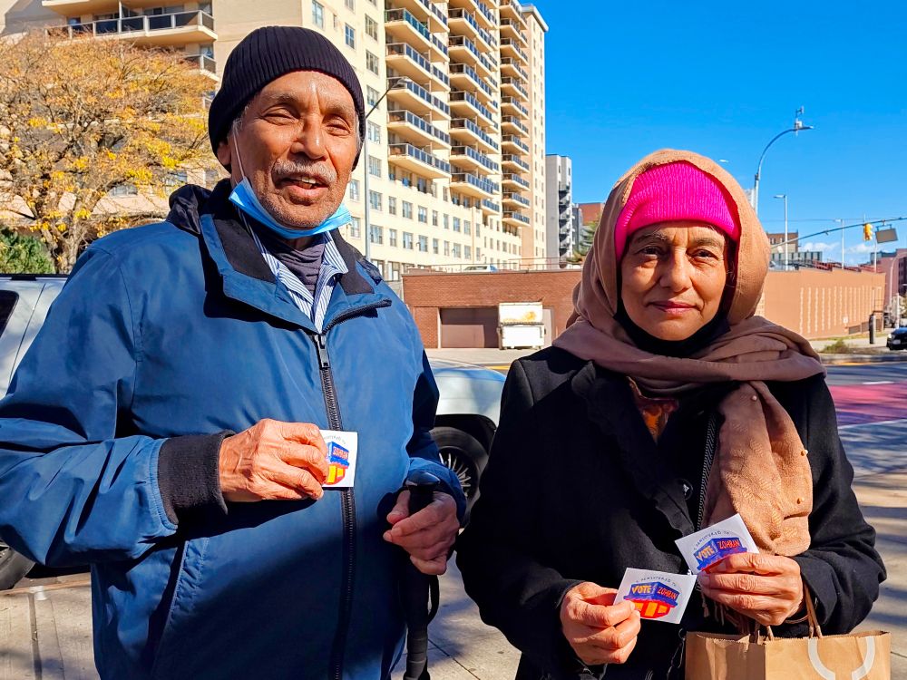 An older South Asian couple happily pose for a photo while holding Zohran campaign stickers. The man, on the left, has lowered his mask and is smiling; the woman wears a bright pink hat and a subtler pink shawl around it while holding two campaign stickers (she wanted an extra one to bring to her relative).