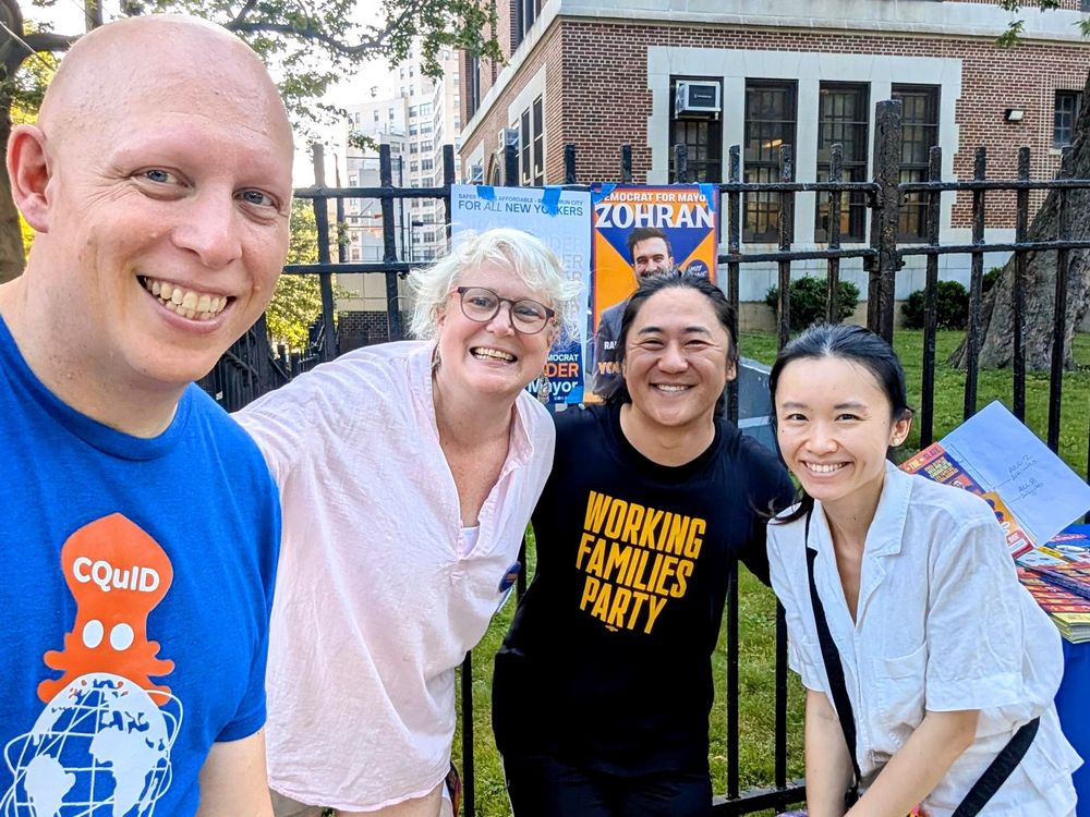 Four volunteers smile for a group photo in front of PS 99 in Kew Gardens, Queens. From left to right: a middle-aged white man, a middle-aged white woman, an East Asian man (of Korean descent), and an East Asian woman (of Chinese descent). The white man is wearing a shirt showing the logo of CQuID [Central Queens Independent Democrats]; the logo consists of an orange-colored squid over a globe that resembles the Unisphere in Flushing Meadows-Corona Park. The East Asian man is wearing a Working Families Party shirt. Visible behind the people are posters of Zohran Mamdani and Brad Lander taped to the fence behind them.