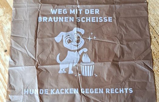 Braun/weisse Hundetüte mit der Aufschrift "Weg mit der braunen Scheisse" "Hunde kacken gegen Rechts" In der Mitte ein Hund der seinen Beutel in einem Mülleimer entsorgt.