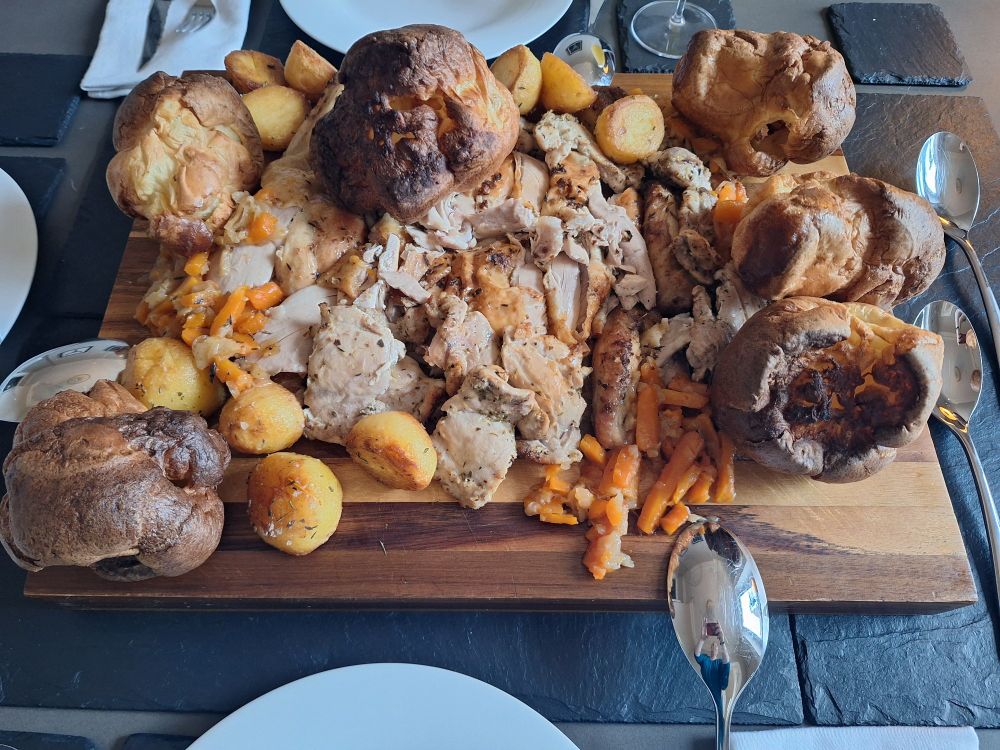 Roast dinner presented on a wooden cutting board, featuring yorkshire pudding, roast chicken, roast potatoes, carrots, and homemade gravy