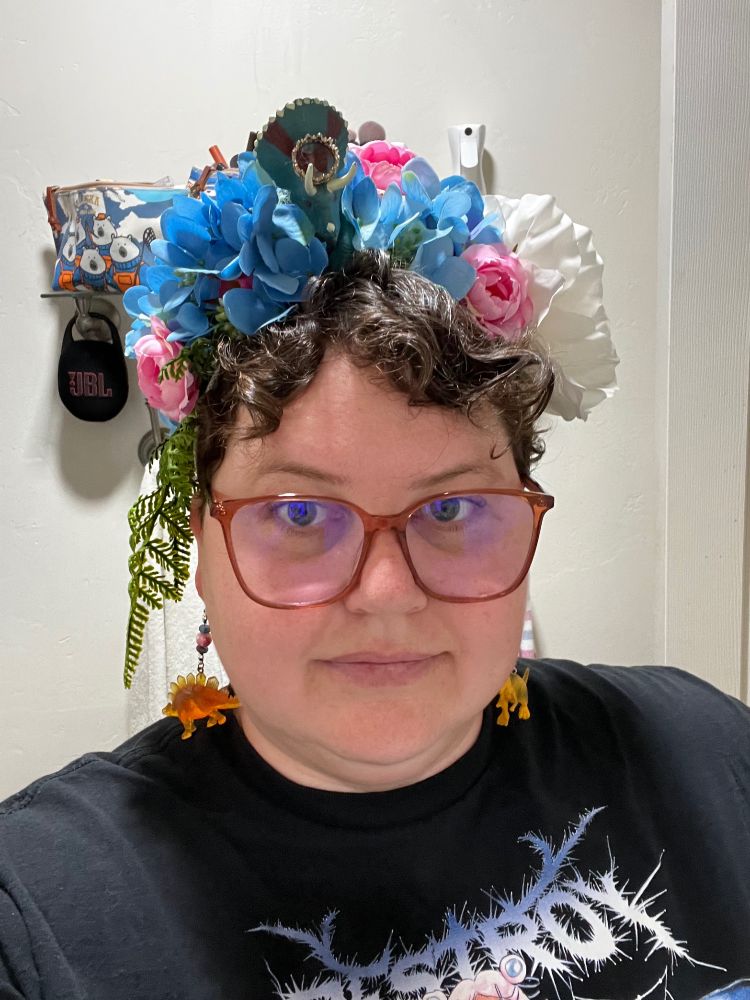 A white person with hazel eyes and dark brown curly hair stands in their bathroom. On their head is a diy dinosaur flower crown. It’s covered with blue hydrangeas with small pink roses, and there are ferns dripping down the left side. On the right side are fluffy white flowers and in the middle is a blue triceratops with a gold crown.