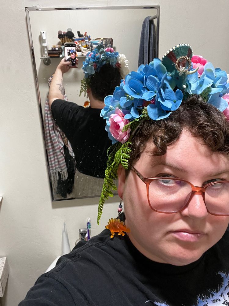 A white person with hazel eyes and dark brown curly hair stands in their bathroom. On their head is a diy dinosaur flower crown. It’s covered with blue hydrangeas with small pink roses, and there are ferns dripping down the left side. On the right side are fluffy white flowers and in the middle is a blue triceratops with a gold crown. They are standing in front of a mirror so you can see the back of the crown as well.