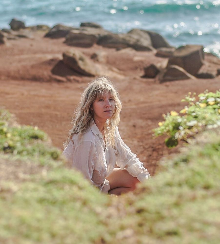 Wavy haired blonde haired woman wearing a white shirt staring at the camera sitting on the beach with grass in the foreground and rocks and ocean in the background 