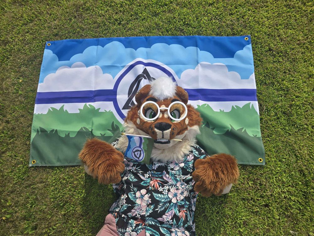 me in my maxi cat fursuit laying on my therian flag design with a small one in my suits mouth, my suit is a brown scottish fold cat with cream neck and chin and white hair with green eyes, the flag is a landscape type scene with 2 grass patterned green stripes, a white cloud pattern stripe and two blue shade cloud stripes, in the middle is a purple stripe with the theta delta in a purple circle