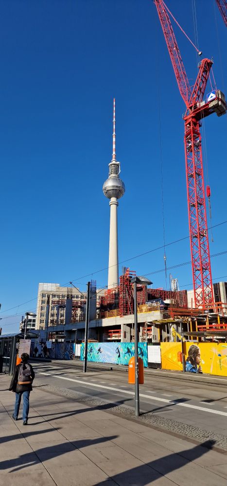 Image of the iconic Berlin TV Tower with bright blue sky behind it, and some cranes undertaking construction.