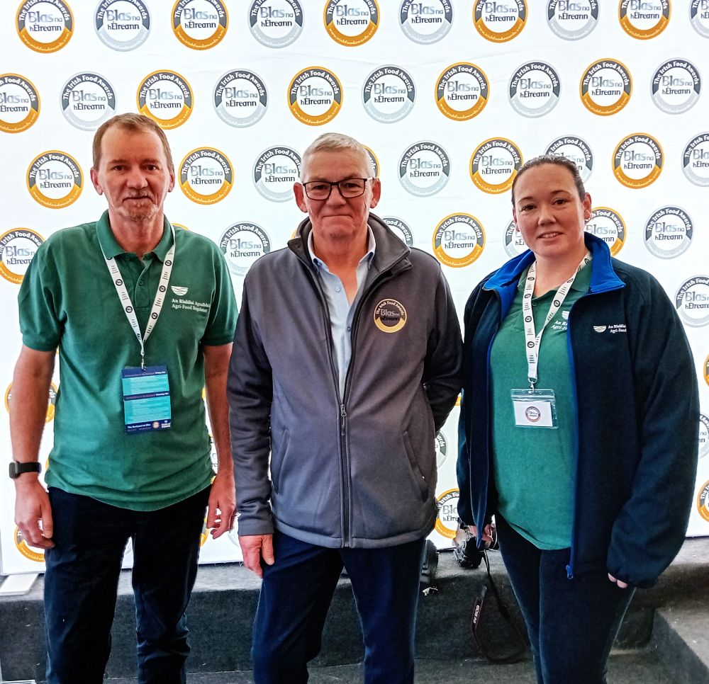 The image shows three people standing in front of a backdrop with the "Blas na hÉireann" Irish Food Awards logo. They are likely at an event related to the awards.