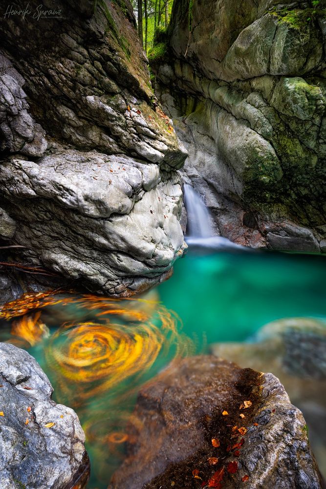 A photo of a gorge in Austria. In the middle ground there is a basin of green water - on the left sine there's a swirl of autumnal leaves blurred by a long exposure to make the movement visible. The rocks in the foreground have some fallen leaves on them too. Higher rock formations in the background surround the small waterfall feeding the basin. Moss is covering part of the rocks.