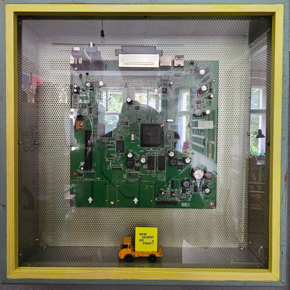 Das Bild zeigt eine grüne Leiterplatte, die in einem quadratischen, durchsichtigen Gehäuse präsentiert wird. Die Leiterplatte ist mit verschiedenen elektronischen Bauteilen bestückt, darunter Widerstände, Kondensatoren und integrierte Schaltungen. In der oberen linken Ecke befindet sich ein großer, schwarzer Chip. An den Rändern der Leiterplatte sind mehrere Anschlussbuchsen sichtbar. Im unteren Bereich des Gehäuses steht ein gelbes Spielzeugauto, das auf einem kleinen, gelben Block platziert ist. Der Block trägt die Aufschrift "WEM GEHÖRT DIE STADT?". Der Rahmen des Gehäuses ist in einem hellen Gelbton gehalten. Im Hintergrund sind unscharf Fenster und Pflanzen zu erkennen. (dieser text wurde mit GPT-4 erzeugt, nur ein ins Auge springender Fehler wurde korrigiert)