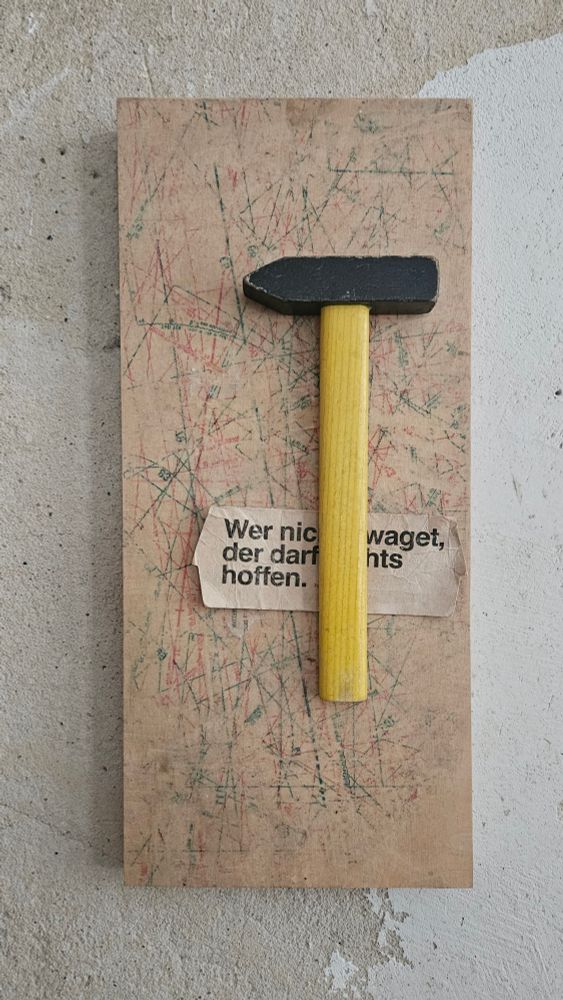 Eine hochkant-rechteckige beige-braune Fläche mit roten und grünen Kratzern, die unregelmäßig verteilt sind. Auf dieser Fläche befindet sich ein Hammer mit einem gelben Holzgriff und einem schwarzen Kopf. Der Hammer ist senkrecht auf dem Papier platziert. Unter dem Hammer ist ein Stück Zeitungspapier sichtbar, auf dem folgender Text zu lesen ist:

Wer nichts waget
der darf nichts
hoffen.

(dieser text wurde mit GPT-4 erzeugt und menschlich redigiert)