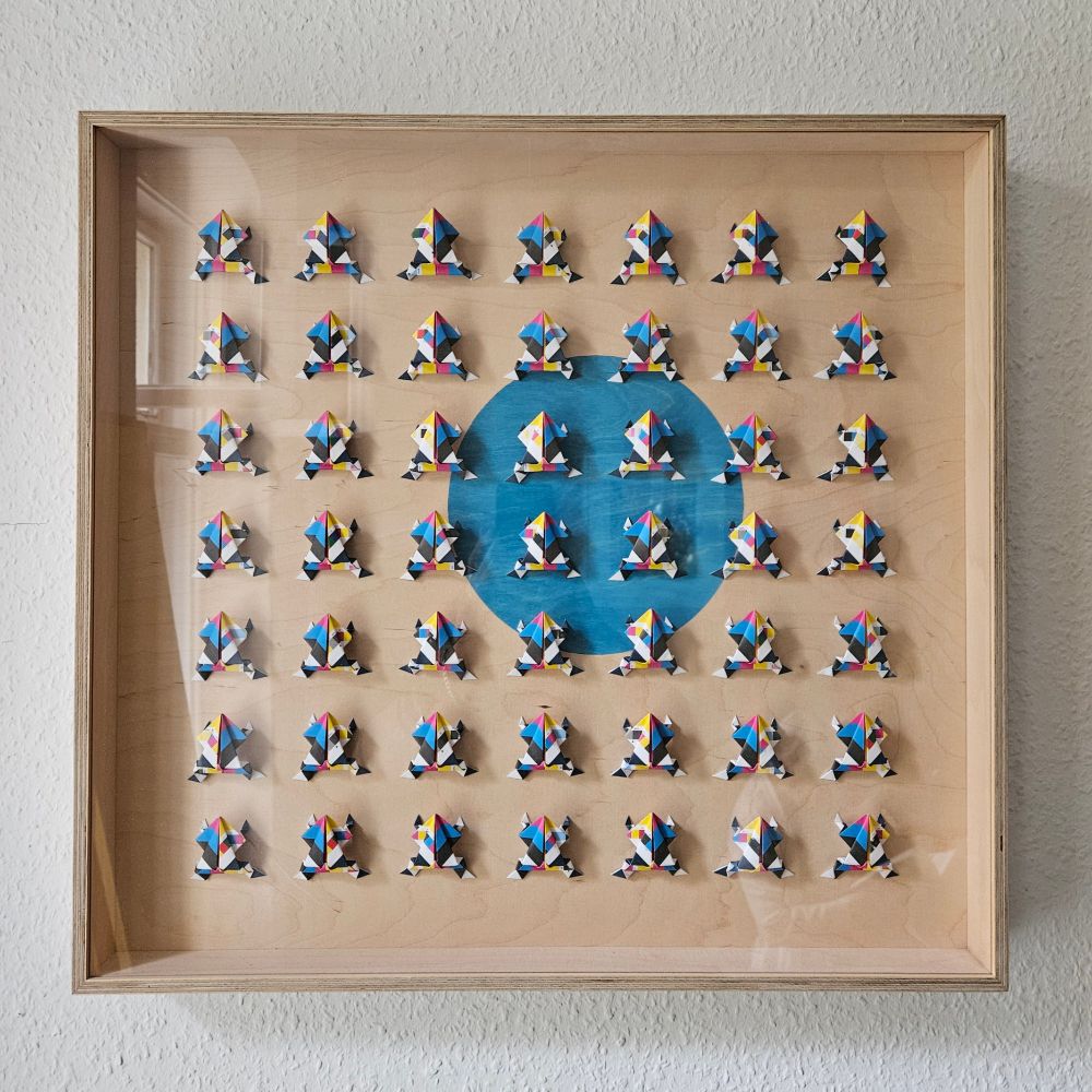 Das Bild zeigt eine quadratische Anordnung von 49 kleinen Papier-Falt-Objekten, die in einem Rahmen präsentiert sind. Jedes Objekt hat die Form eines stilisierten Froschs, der kubistisch anmutend aus verschiedenen geometrischen Formen zusammengesetzt ist. Die Frösche sind geometrisch unterteilt in Rot, Blau, Gelb, Schwarz und Weiß gehalten. Auf den aller ersten Blick scheinen sich die Musterungen zu gleichen, auf den 2. Blick scheint sich keine exakt zu wiederholen.
In der Mitte des Rahmens befindet sich ein großer blauer Kreis, der einen Kontrast zu den bunten Fröschen bildet. Der Hintergrund des Rahmens ist aus hellem Holz gefertigt, was die Frösche hervorhebt. Die gesamte Komposition ist symmetrisch und gleichmäßig verteilt, die Frösche bilden ein reglemäßiges Raster.
(dieser text wurde mit GPT-4 erzeugt und menschlich redigiert)