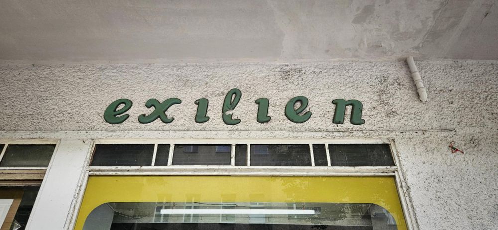 das bild zeigt eine wand mit einem beschrifteten schild. das schild trägt die aufschrift "exilien" in grüner schrift. die buchstaben sind in einer geschwungenen, leicht verspielten schriftart gestaltet. der hintergrund der wand ist grau und weist eine raue oberfläche auf. unter dem schild sind fenster zu sehen, die teilweise mit gelbem material umrahmt sind. die fenster sind geschlossen und reflektieren das licht. die gesamte szene wirkt schlicht und unauffällig. (dieser text wurde mit GBT-4 erzeugt und nicht lektoriert)