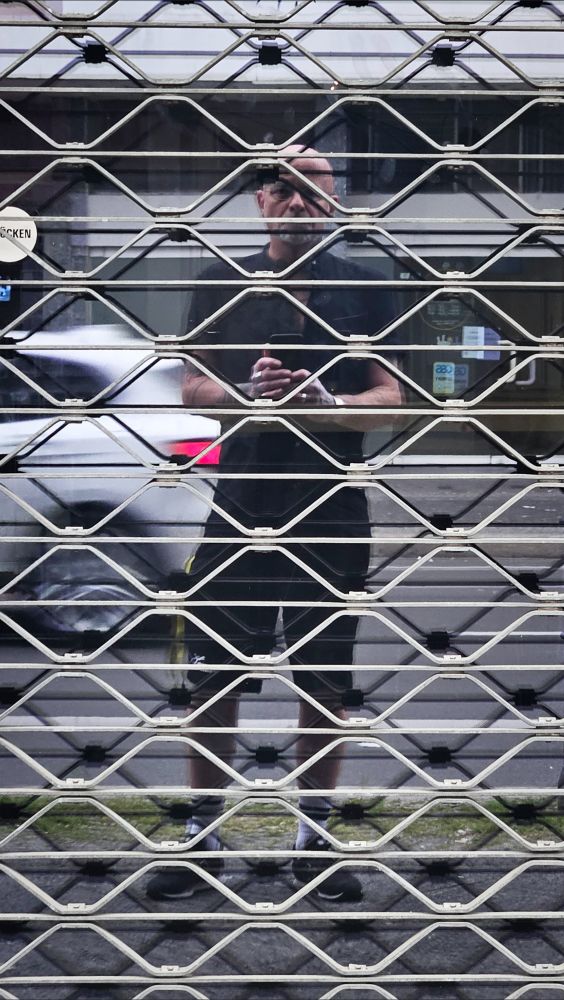 das spiegelbild einer männlichen person in der vergitterten eingangstür eines geschlossenen geschäfts. sie trägt ein kurzärmliges schwarzes hemd und schwarze shorts, hat tätowierte unterame und hände mit einigen silberringen, hält das smartphone in der hand und blickt auf seine reflexion. im hintergrund ist bewegungsunscharf ein vorbeifahrendes autos und eine straße zu sehen. (text erstellt mithilfe von GPT-4o mini von duckduckgo)