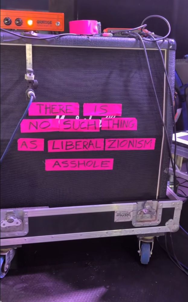 a four speaker amplifier cabinet with black grill sitting on a road case with wheels draped in instrument cables and audio equipment. strips of bright pink duct tape on the grill are lettered with black marker to say THERE IS NO SUCH THING AS LIBERAL ZIONISM ASSHOLE 