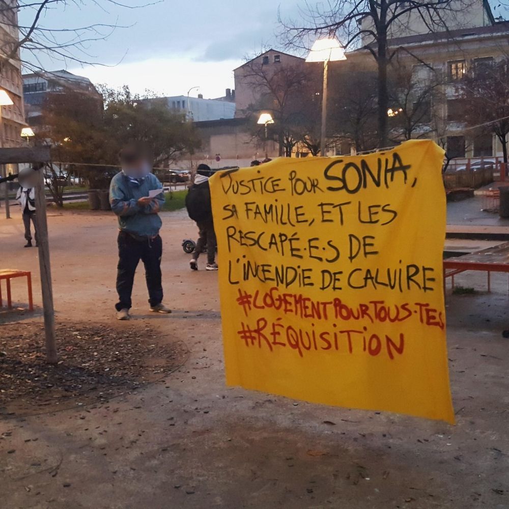 photographie de la conférence de presse des soutiens du squat denuzières à lyon place mazagran. entre deux arbres, une banderoles jaune accrochée, écrit : "justice pour sonia, sa famille et les respacapé.e.s de l'incendie de caluire #logemementpourtous.tes #réquisition"