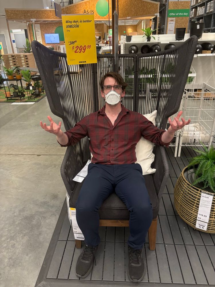 A masc person with short brown hair, clear plastic glasses, and a Flo mask sits in a ridiculous black outdoor chair with oversized wings comprised of black elastic. For some reason.

He makes an evil frown and raises his hands in a clawed, dramatic “you walked into my trap” sort of way.