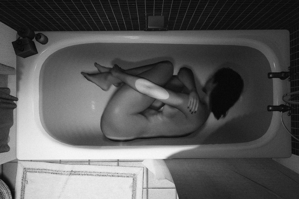 Blank and white picture showing an overhead view of a naked woman curled into a fetal position beneath the water of a white bathtub.