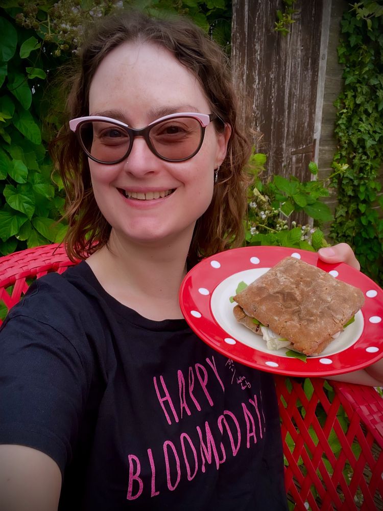 A smiling middle aged white woman with wavy brown hair and glasses holding up a plate with a sandwich on it. Her t-shirt says Happy Bloomsday!