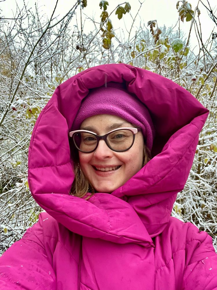 Cleo dressed in a shocking pink puffer coat, hood pulled up over a matching pick fleecy hat. She’s smiling and standing in front of some snow covered tree branches.