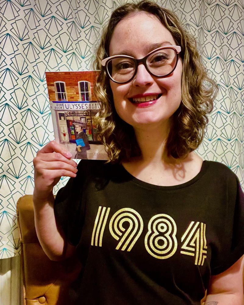Photo of Cleo - a 40 year old woman with curly light brown hair wearing funky black and pink glasses and a black t-shirt with ‘1984’ written on it in gold. She is holding up a birthday card with a picture of James Joyce on it.