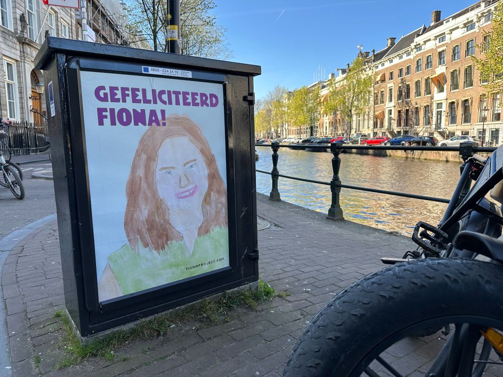 A poster that reads 'GEFELICITEERD [aka happy birthday] FIONA' with a watercolor profile of a woman, next to a canal in Amsterdam.