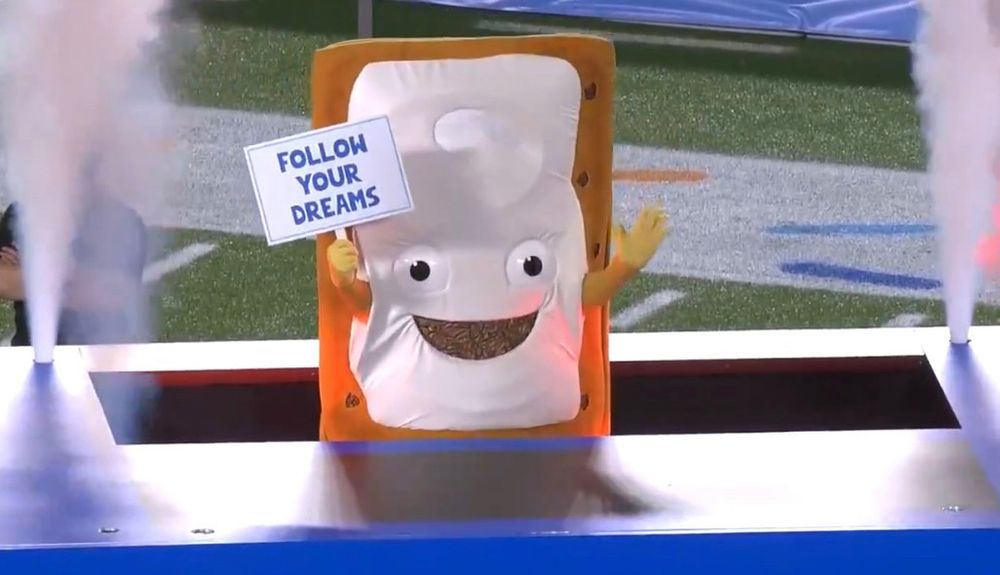 Someone in a large pop tart suit with a sign reading follow your dreams being lowered into a toaster on a football field