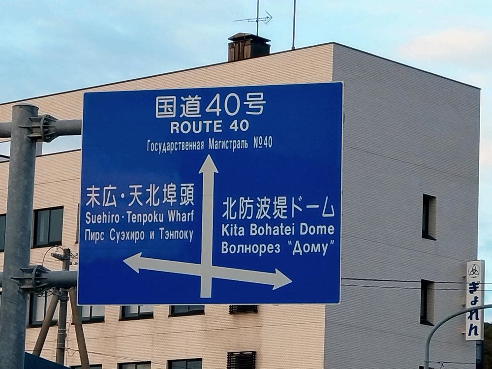 国道40号の青い道路標識。行き先は左が末広・天北埠頭、直進が北防波堤ドーム。日本語、英語、ロシア語で表記。背景に建物。