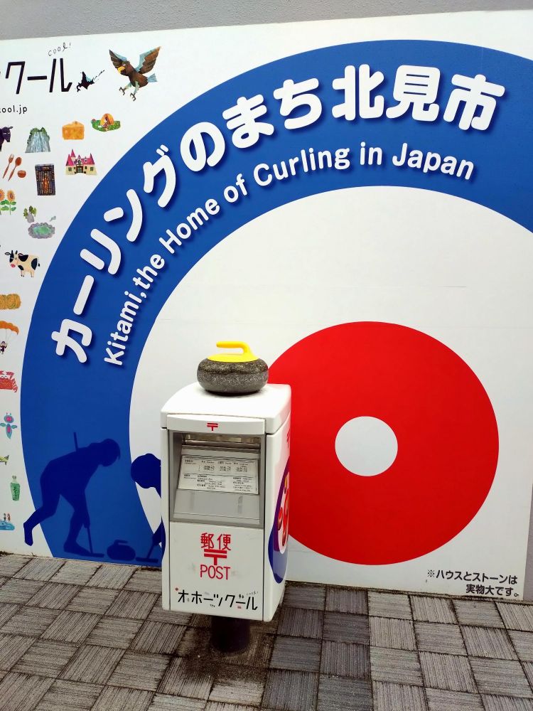 カーリングのストーンが載った郵便ポストと、背景に「カーリングのまち北見市 Kitami, the Home of Curling in Japan」と書かれた看板。ポストには「郵便 POST オホーツクメール」と記載。