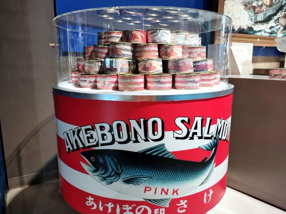 缶詰「AKEBONO SALMON PINK あけぼの さけ」の大型ディスプレイと、内部に積み上げられた様々な種類の缶詰が展示されている様子。