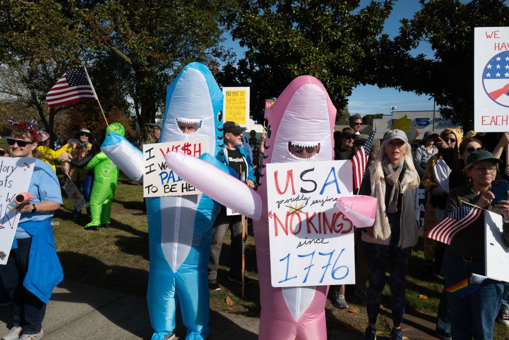 Two people in inflatable shark costumes. Signs read: "I wish I was paid to be here" and "USA proudly saying no kings since 1776"