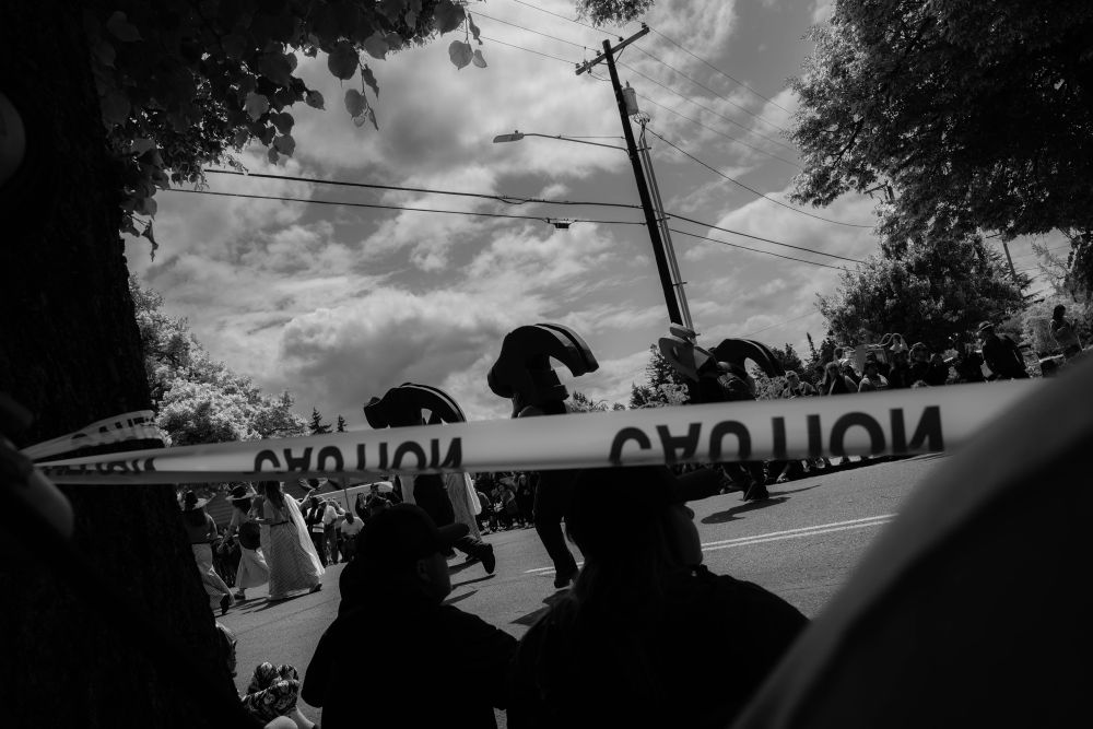 A high-contrast black and white photo of a street festival with caution tape in the foreground and the silhouette of two people sitting on the curb next to a tree. Four figures dressed as large hammers retreat ln the street in the distance. 