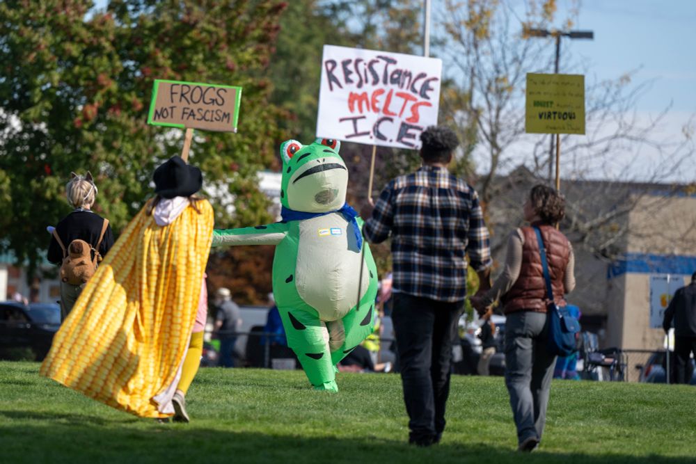 Three people walk away from the camera as an inflatable frog dances in the park. Two signs read "FROGS NOT FASCISM," "RESISTANCE MELTS ICE"