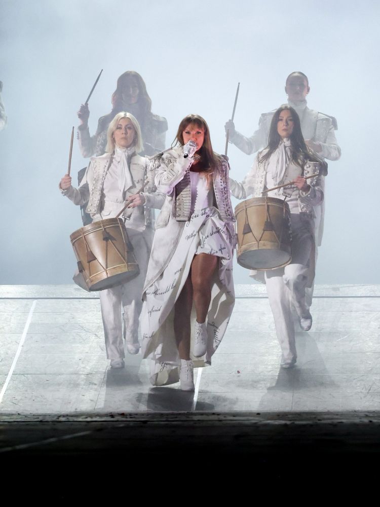 Taylor Swift in a white military jacket and white textured dress, marching while singing, leading drummers during The Smallest Man who ever lived , during the Eras Tour
