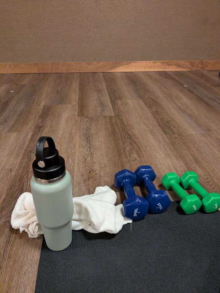 Picture of a green insulated water bottle by a towel and two sets of weights at the top of a yoga mat. The mat is on a wood floor with a brown wall in front of it.