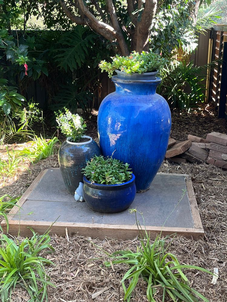 Three blue glazed pots on a grey ceramic plinth. The garden around it has green strappy leaved plants and light brown mulch. 