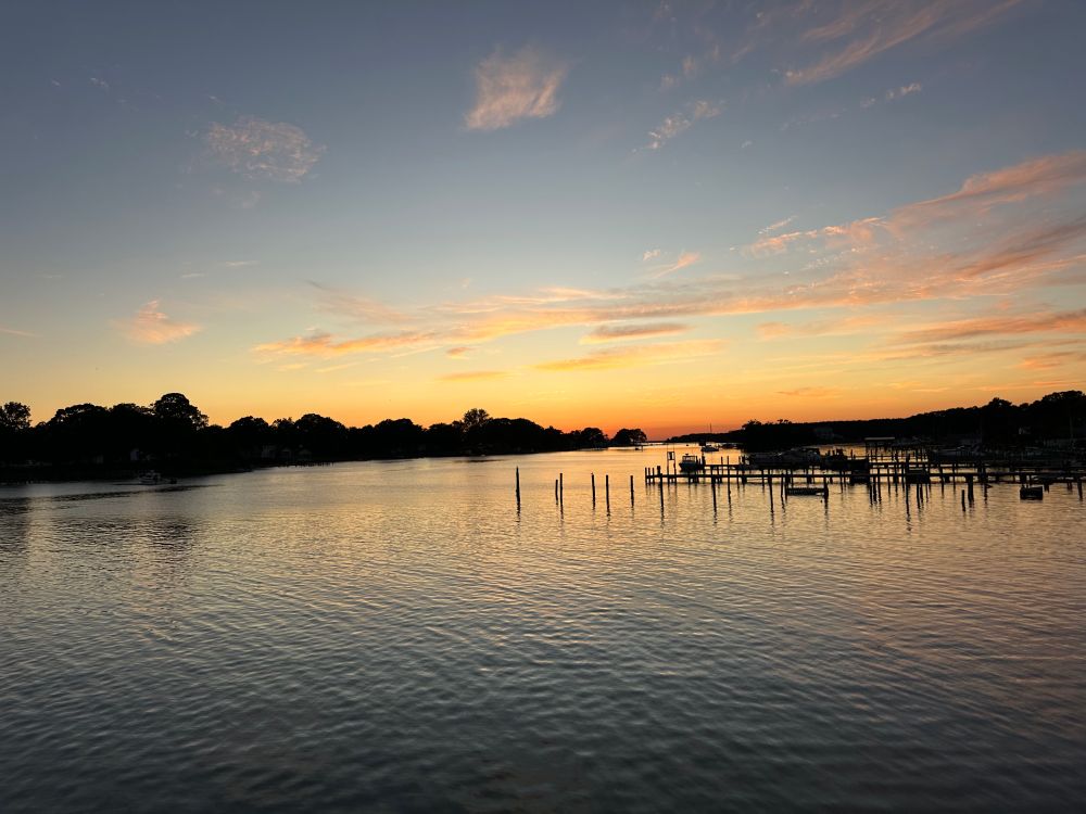 Picture of a waterway in Cobb Island, MD at sunset. The sky and clouds are gradients of orange to blue which is reflected in the rippling water. There are trees on both sides of the waterway in silhouette along with boat piers.