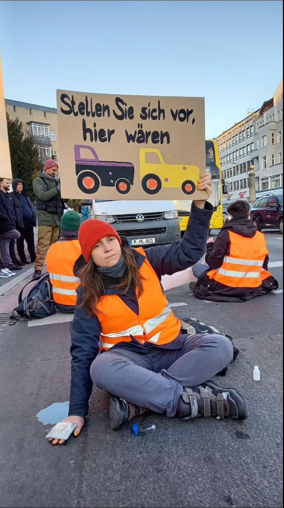 Picture of a Last Generation protestor in Germany whose one hand is glued to the street while she holds a sign that says ‘imagine there were tractors here’ (the word tractor is not spelled out, but two tractors are painted on the sign). 