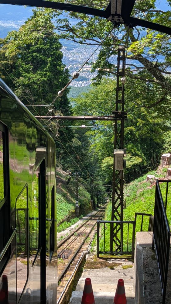 A lush green forest lies on a steep hill with a train track. There is a cable car in the left of frame with the reflection of the beautiful green grass. In the distance, the skyline of Kyoto can be seen.