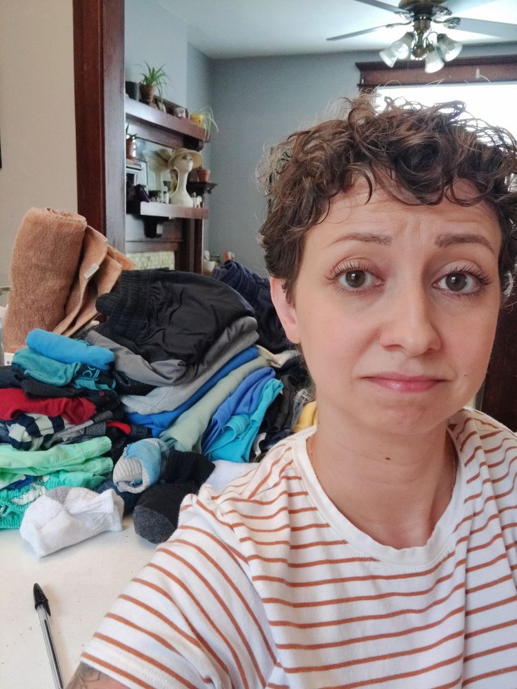 A sad looking woman with brown curly hair in a white and orange striped shirt sits in front of a pile of folded kid's clothes.