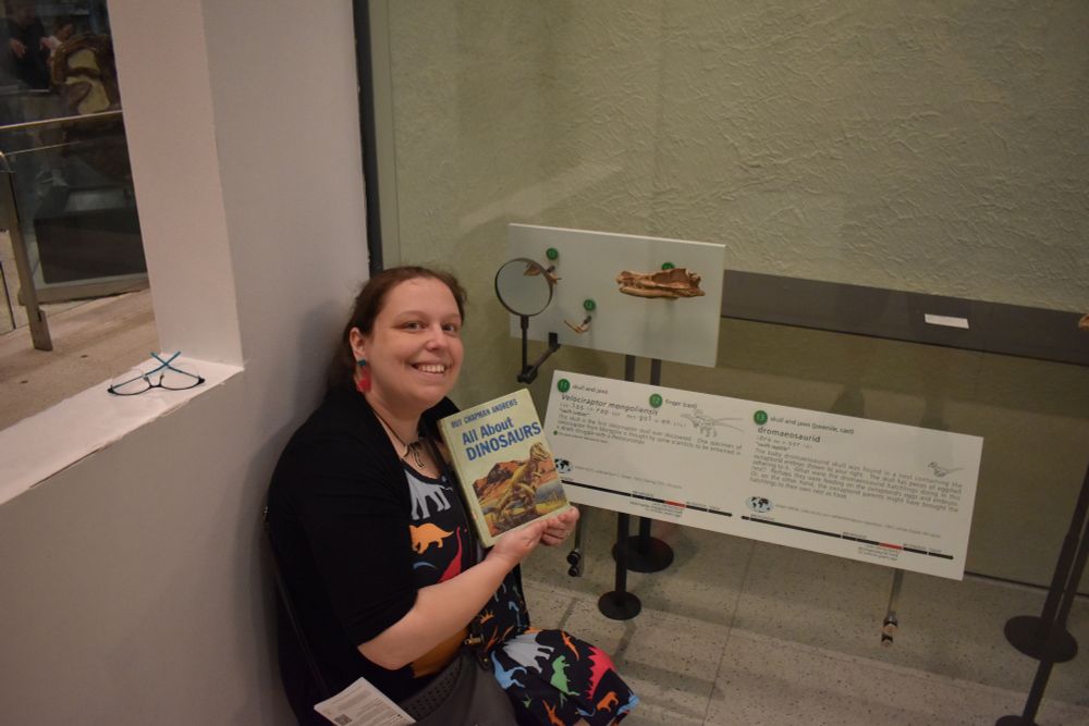 An elated paleontologist wearing a neon and black-colored dinosaur dress with pink dinosaur earrings sitting next to three Mongolian dinosaur specimens behind a glass case. The paleontologist is holding "All About Dinosaurs" by Roy Chapman Andrews, who was involved in the 1923 Central Asiatic Expedition that recovered the Velociraptor skull in the case.