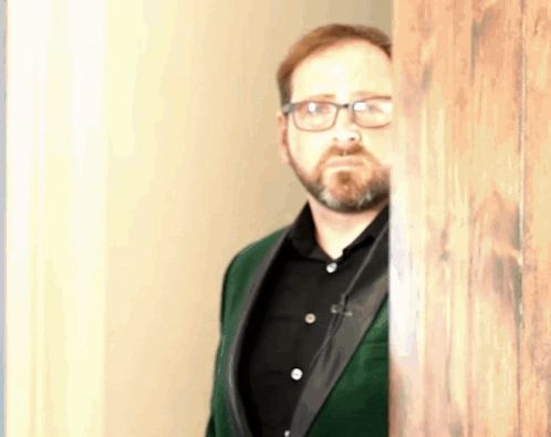 a man with glasses and a beard is peeking out from behind a wooden door