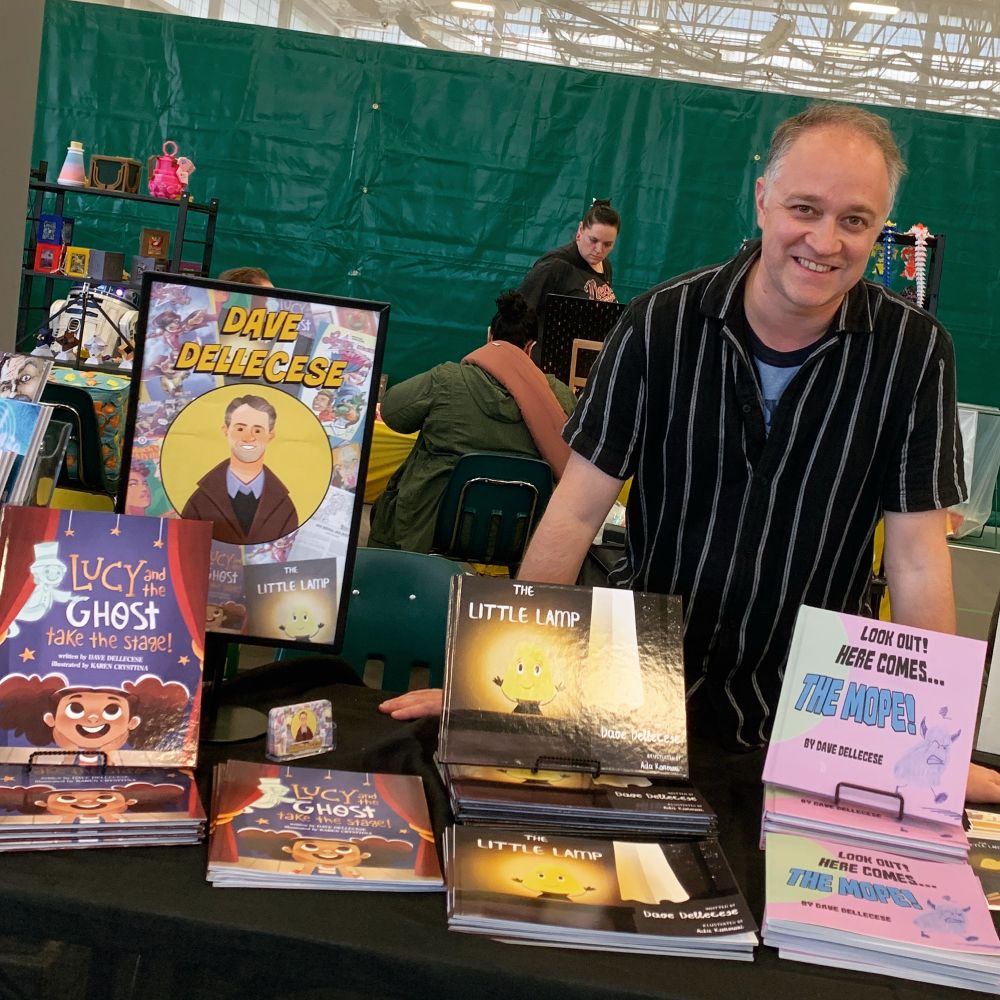 Dave, wearing a black and white striped camp shirt, leans on a table where various children’s picture books sit.