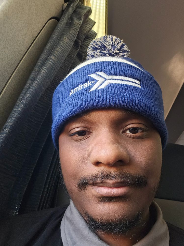 Handsome young black man sitting on a train wearing a blue and white winter hat with a white puff on top and the retro Amtrak logo on the rim