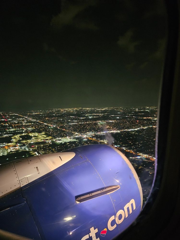 A nighttime view out of a plane window descending into a city, with the plane's blueish purplish engine and the city lights following major streets in a grid in the background under a night sky with wispy clouds reflecting the light from below
