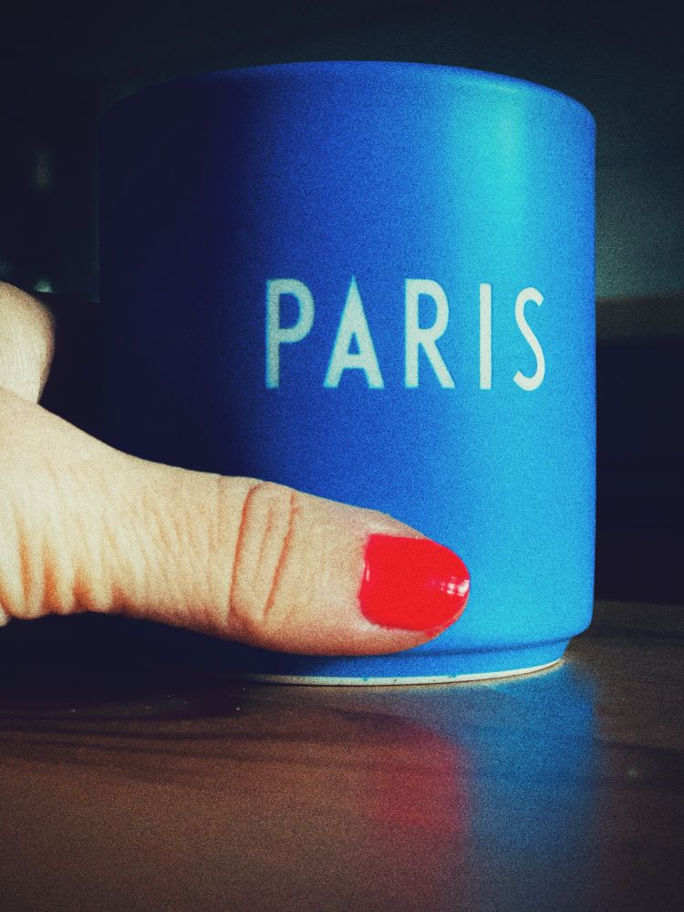 Blaue Tasse mit weißem Schriftzug „Paris“, links eine Hand mit rotem Nagellack, die die Tasse hält.