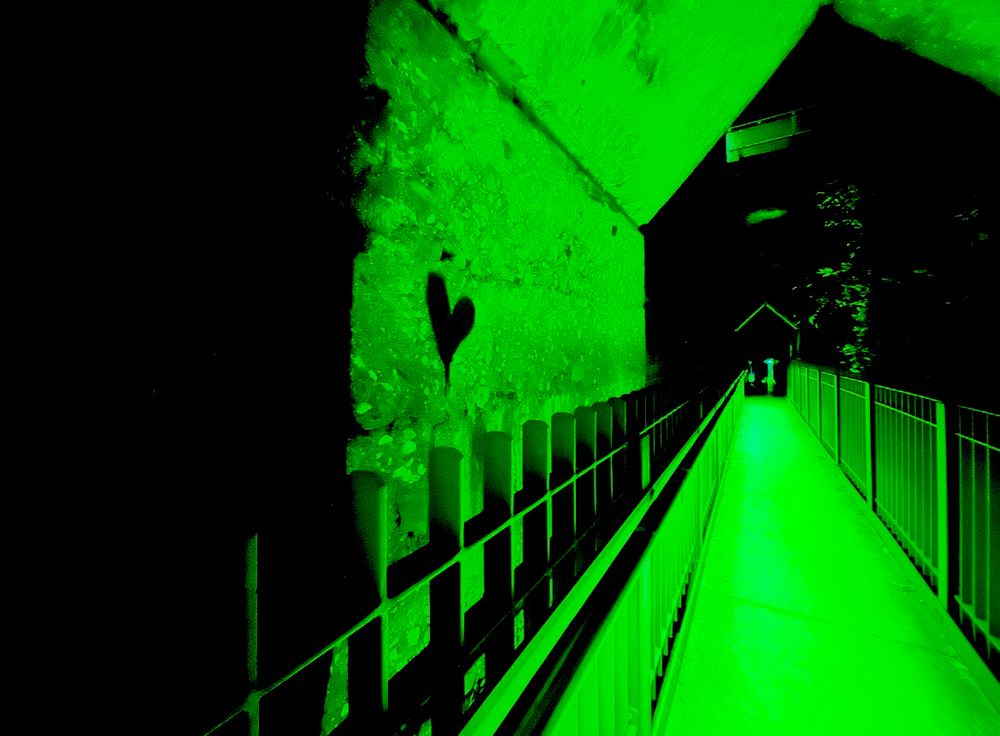 Gang in altem Fabrikgelände, in grünes Licht getaucht,an sieht nur einen Weg mit Zaun. Links im Bild eine Wand mit einem Herz darauf.