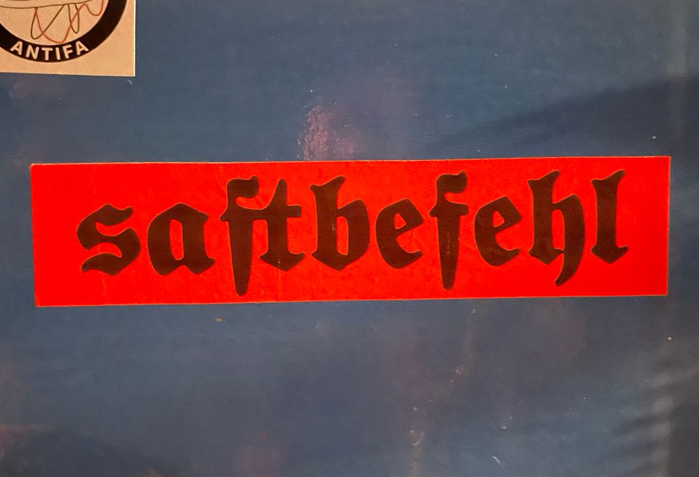 Orangefarbener Sticker mit dem schwarzen Fraktalschriftzug „saftbefehl“.