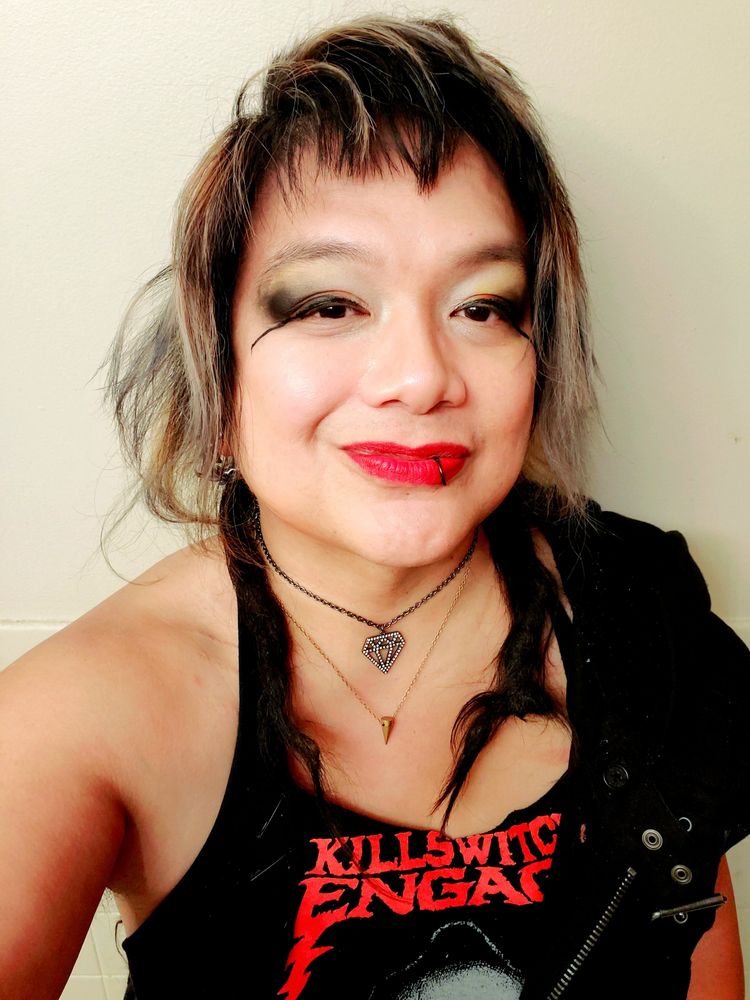 Kei, a tan heavy set Filipunx Trans Woman, standing against a light yellow wall and shot from chest up, wearing a black KILLSWOTCH ENGAGE tee cut and pinned into a crop halter tank with a black front zip buckle best worn on left shoulder, brass cone and die-cut diamond bling necklaces, red lipcolor and white to yellow with black dust outer eyeshadow amd extended puppy eyeliner, makeup faded and worn from a long day at work, short black dread tips forward on shoulders and black and bleached side wispys and choppy bangs framing face, She's got a rare smile because She just found out that Trans Appreciation Friday is Back! ^___^✨️