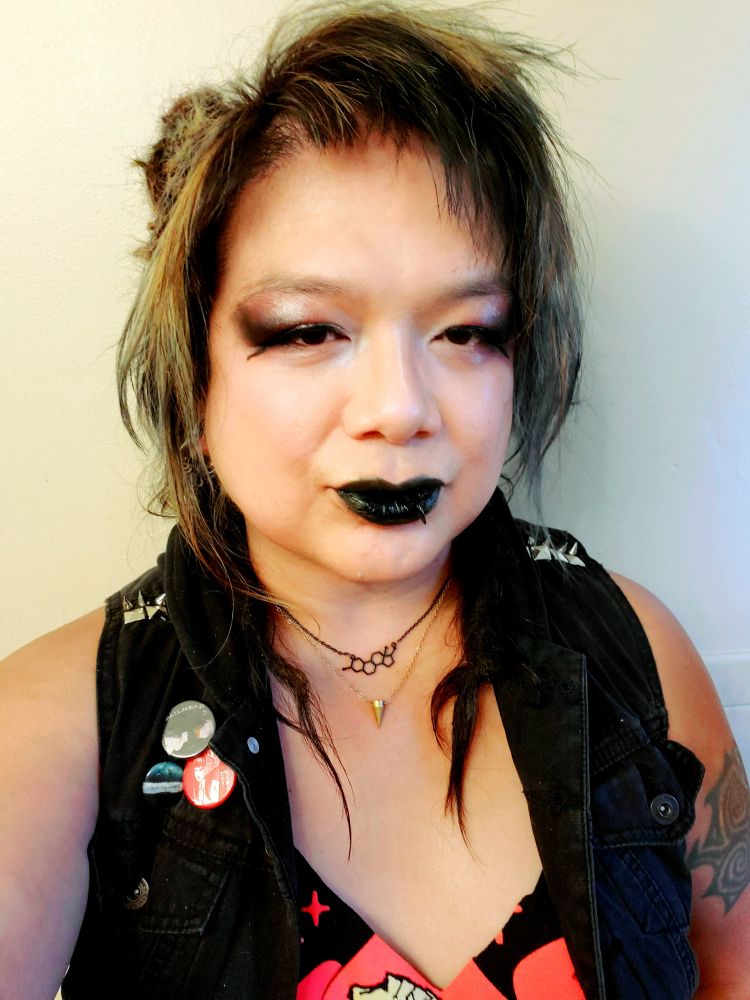 Kei chest up in a black with red print deep v-neck cut tee under a black denim vest with spikes and studs on shoulders and pins above Her right front pocket, MagicBullet and Estrogen chain necklaces, Ling dreads back and short loose dreads forward on shoulders with bangs and wispys brushed down, black lipstain with white to red and raisin with black dust outer eyeshadow with silver accent and puppy eyeliner, room vanity lighting